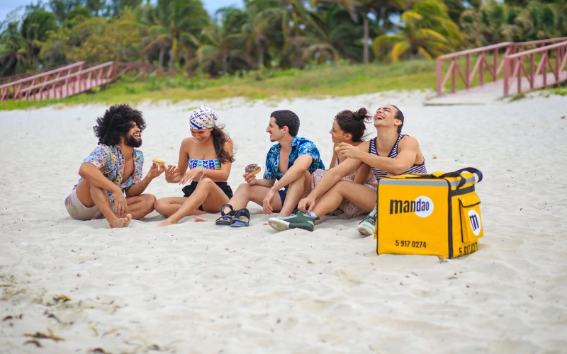 Amigos jÃ³venes riendo en la arena de la playa Varadero con la mochila amarilla del servicio de delivery de Mandao en Matanzas, Cuba. 